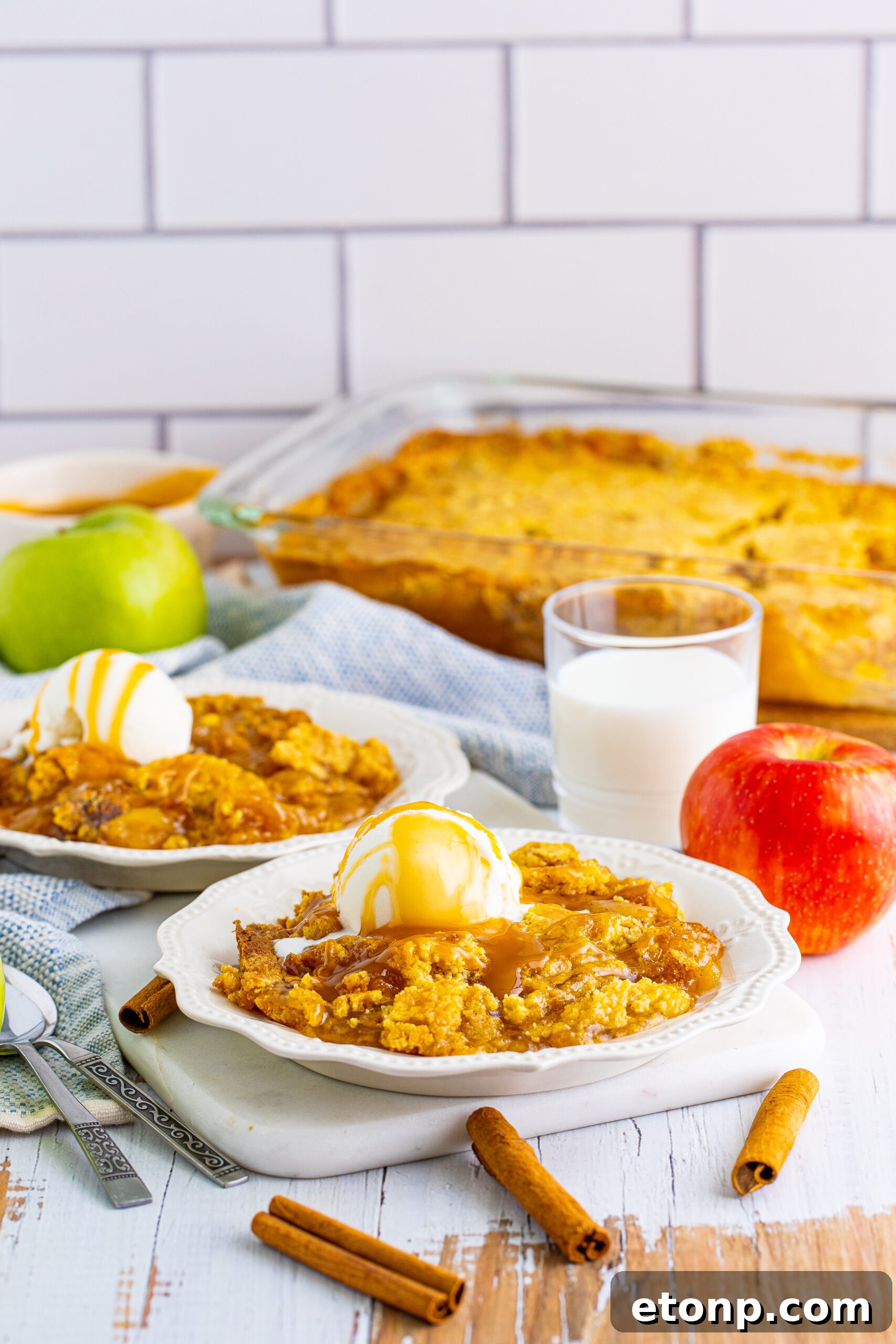 Caramel apple dump cake on two white plates with cinnamon sticks and apples in the background.