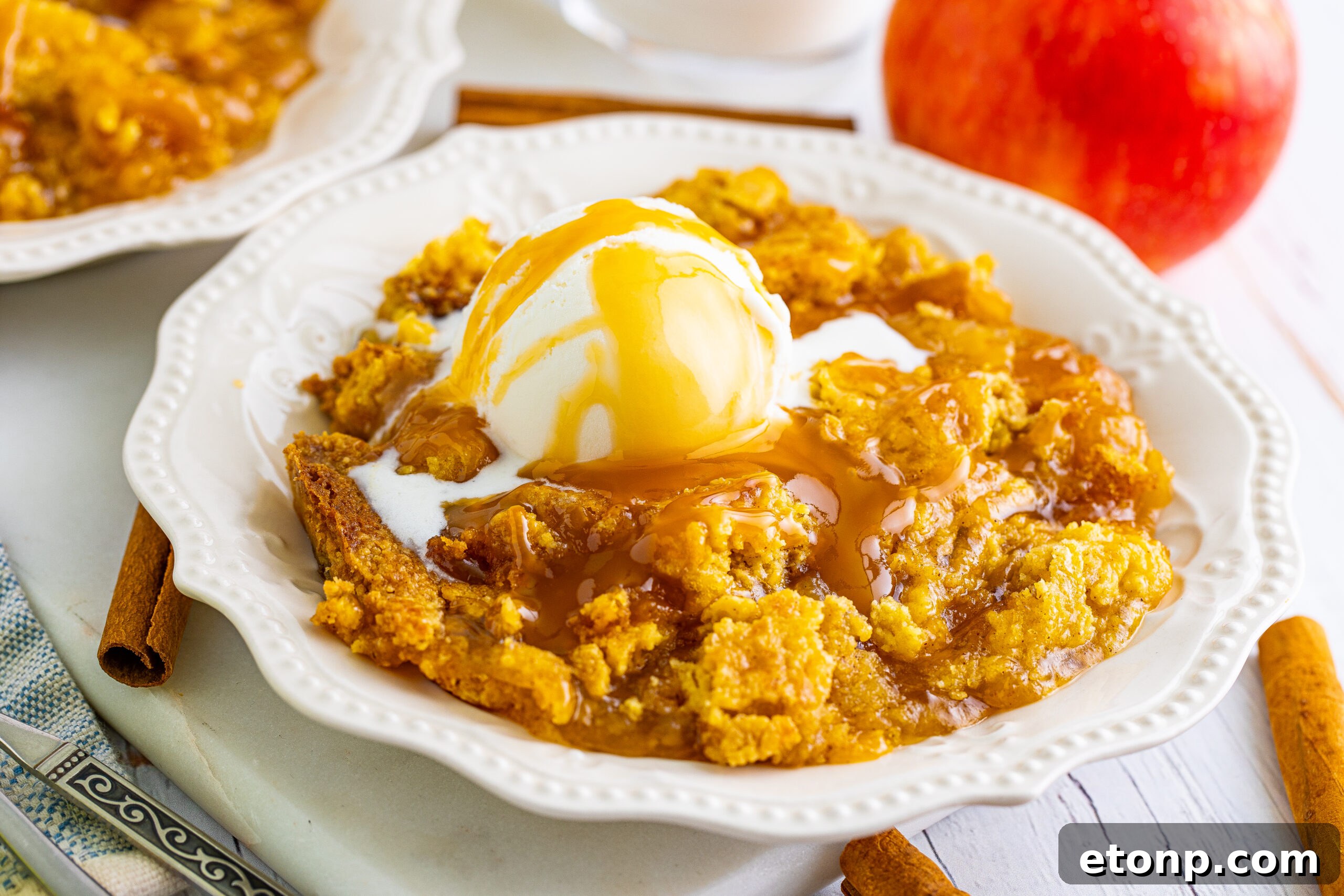 Close up shot of caramel apple dump cake on two white plates with cinnamon sticks and apples in the background.