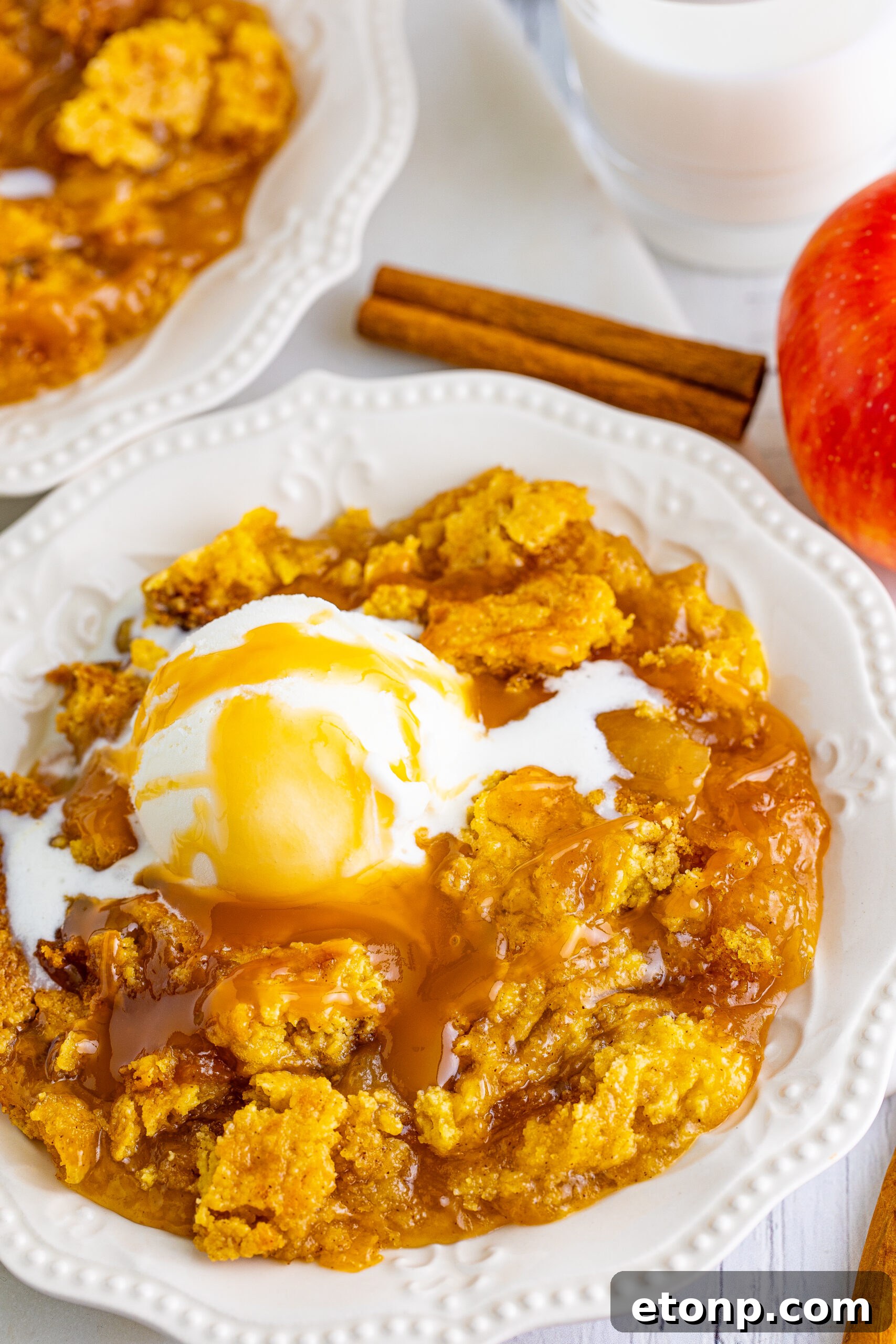 Close up shot of caramel apple dump cake on two white plates with cinnamon sticks and apples in the background.