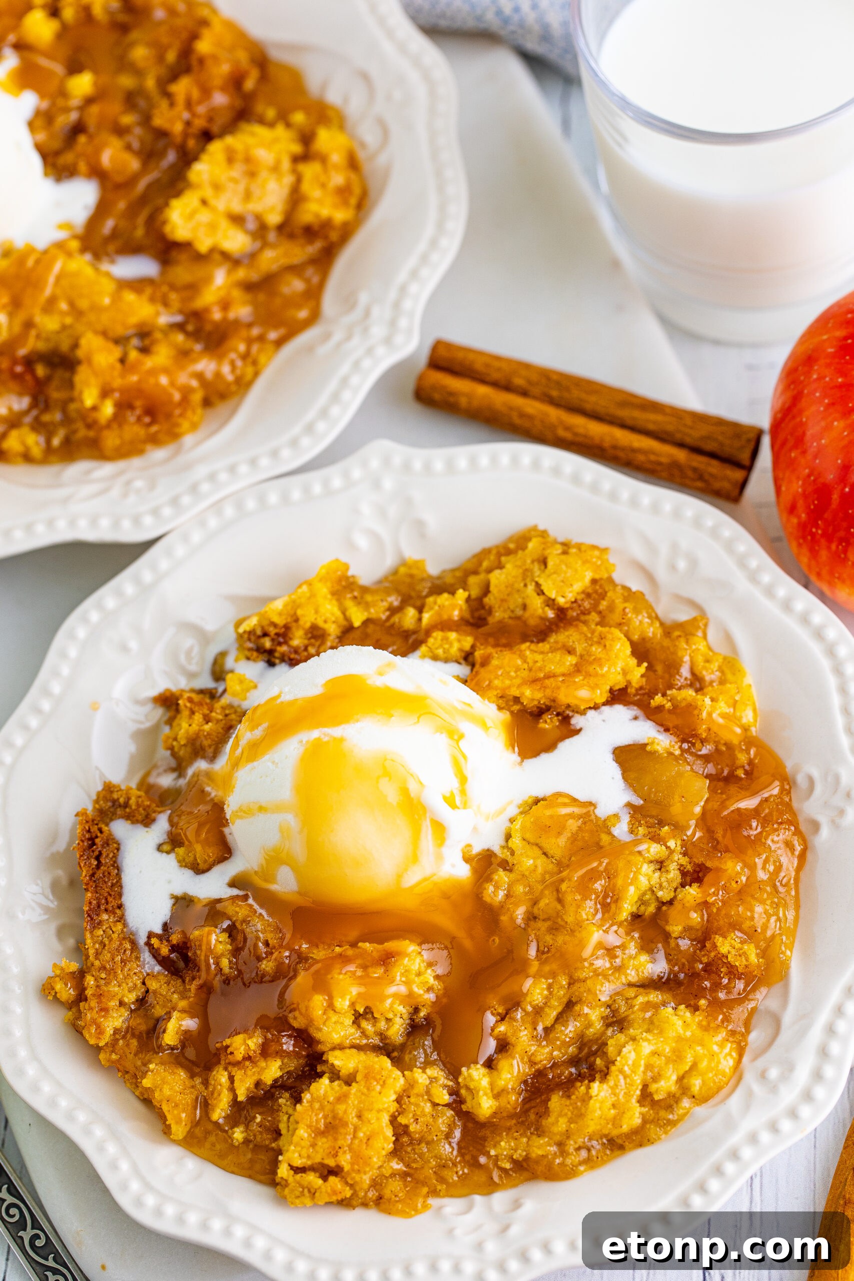 Close up shot of caramel apple dump cake on two white plates with cinnamon sticks and apples in the background.